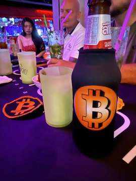 A Bitcoin-branded bar features bottles and napkins emblazoned with the orange "₿" Bitcoin logo on a table, where a man and a woman are seated enjoying drinks, reflecting the venue's crypto-themed atmosphere described in the caption.