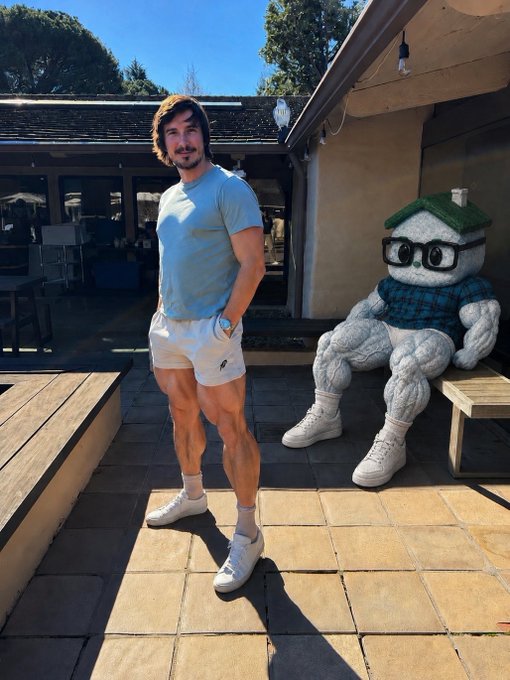 A young man in a light blue t-shirt and white shorts stands on a sunlit patio next to a large, muscular mascot statue wearing glasses, a green beanie, and a plaid sweater, which appears to represent the Robinhood brand referenced in the caption. The image humorously illustrates the post's joke about "Robinhood" getting "legs" by literally depicting the company's mascot with sculpted, muscular legs.