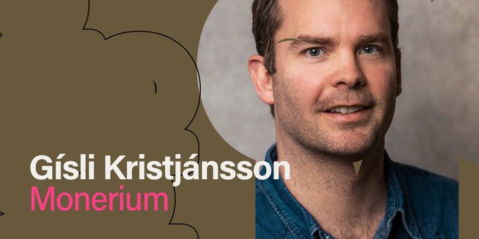 The image features a man wearing a blue shirt with white text that reads "Gisli Kristjansson Monerium". The background of the image is a light brown color, which contrasts nicely with the man's blue shirt. The text on the image is in white and pink colors, making it stand out against the brown background.