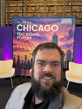 A bearded man in a gray jacket smiles at the camera in front of a 'CHICAGO TDC Signal Forum' sign dated 10.08.26, hosted by the Digital Chamber.