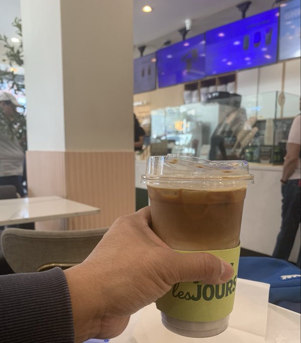 A hand holds an iced latte in a yellow "les JOURS" cup at a coffee shop, capturing a relaxed Monday morning break before diving into crypto lessons.