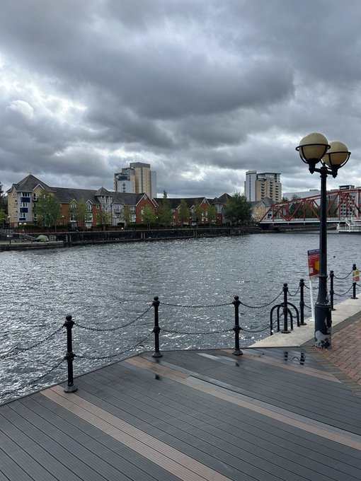 A cloudy day overlooks a waterfront boardwalk with chain-link railings, a decorative streetlamp, and townhouses across the water.