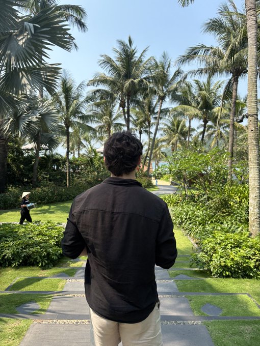 A man with dark curly hair wearing a black shirt and light shorts stands with his back to the camera on a garden pathway lined with palm trees and tropical greenery.