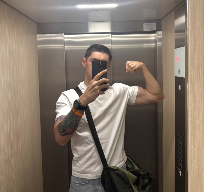 A mirror selfie in an elevator of a muscular man in a white t-shirt flexing his bicep, holding a phone, with visible tattoos on his arm.