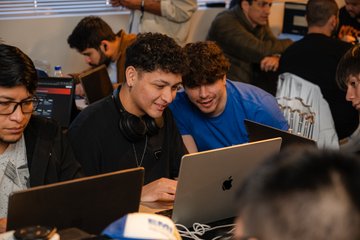 Two young men smile while looking at a laptop together at what appears to be a busy gaming or tech event.