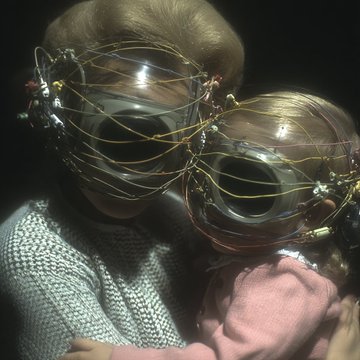 Two dolls wearing intricate wire and bead headpieces, representing the sold NFT artworks "motherform #26" and "metaformatika #42" by the artist, displayed against a dark background.