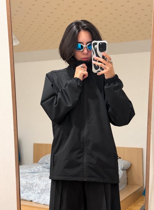 A mirror selfie of a person with short dark hair wearing blue sunglasses and a black hooded jacket, holding up a phone with a black and white case in a bedroom setting.