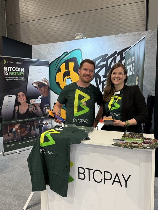 The image shows a man and woman standing behind a table, wearing green shirts with the letters "BTCPAY" printed on them. They are positioned next to a booth displaying their shirts for sale. The booth also features a sign that reads "We're waiting you at #49! Come say hi". This suggests that they are promoting their shirts and possibly attending an event or gathering related to Bitcoin payments, such as a cryptocurrency conference or fair.