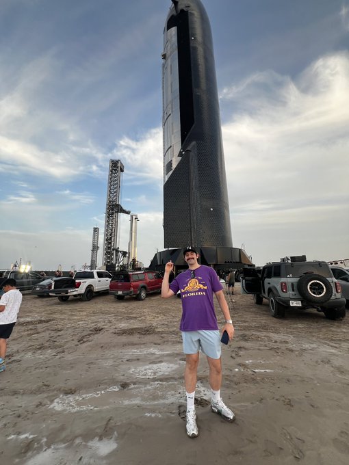 A young man in a purple 'FLORIDA' t-shirt and light blue shorts posing in front of SpaceX's Starship rocket at the launch site.