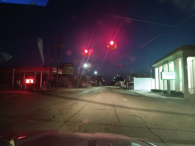 A nighttime view through a car windshield showing red traffic lights at an intersection with a 'BEST IN THE WEST' sign visible on the left and a JTH storefront.