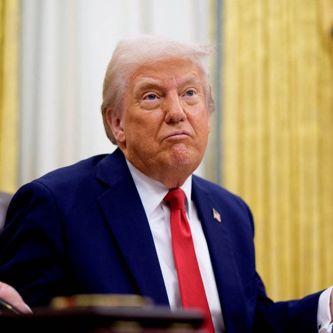 President Donald Trump, wearing a navy blue suit with a red tie, is seated in what appears to be the Oval Office, looking off to the side with a serious expression.