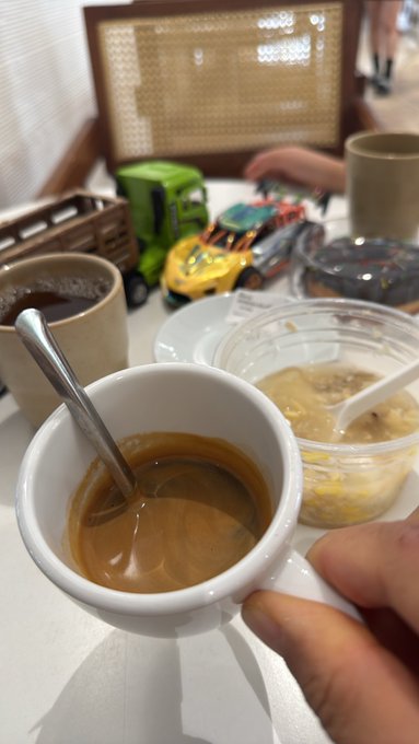 A hand holding a white espresso cup filled with coffee, photographed in what appears to be a café setting, with other drinks, a dessert, and a child playing with toys visible in the background.