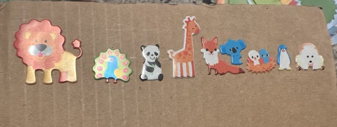 A row of cute animal stickers or cutouts, including a lion, peacock, panda, giraffe, fox, koala, penguins, and a hedgehog, arranged on a cardboard surface.
