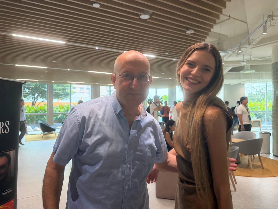 An older bald man in a blue shirt standing next to a young smiling woman with long blonde hair in what appears to be a modern indoor public space.