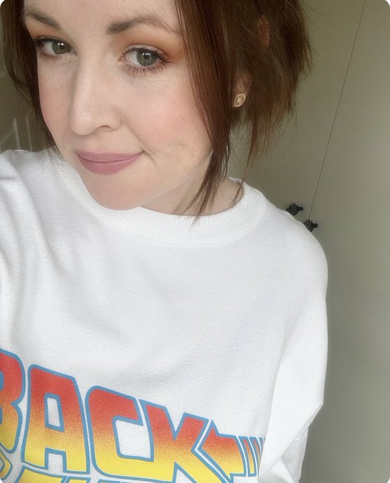 Close-up selfie of a woman with reddish-brown hair wearing a white sweatshirt with colorful 'BACK' lettering.