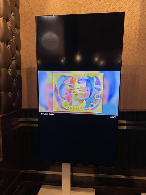 A digital display screen showing a colorful abstract painting by Shakifar NFTTM, set against a tufted brown leather headboard in a warmly lit room.