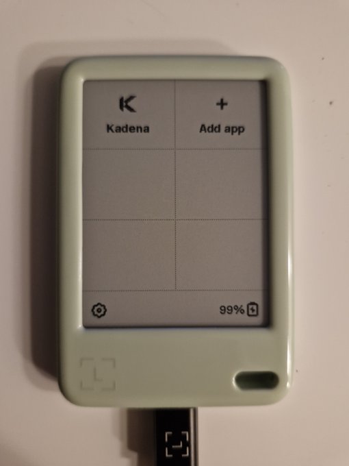 A small mint-green e-ink device displaying two app tiles labeled 'Kadena' and 'Add app', with a battery indicator showing 99% and a charging cable plugged into the bottom.