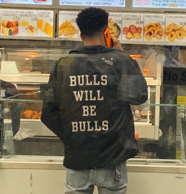 A man wearing a black denim jacket with "BULLS WILL BE BULLS" printed on the back stands at a fast-food counter while talking on an orange phone, with menu items like pizza rolls ($11.75), mac & cheese mashed potato ($2.00), onion rings ($2.50), and chicken rings ($14.99) visible above him. The "BULLS WILL BE BULLS" slogan mirrors the post's persistent "we're gonna keep showing up" energy, playfully equating relentless shilling to a stubborn bull mindset—eventually wearing you down into buying $ANSEM.