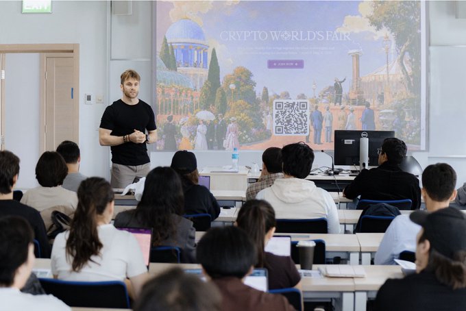A bearded presenter in a black t-shirt stands at the front of a modern lecture hall, addressing an audience of attentive students seated at desks, with the projected slide behind him reading "CRYPTO WORLD'S FAIR" alongside a scenic illustration and a QR code. The scene captures the speaker's talk at Nazarbayev University, organized by Superteam Kazakhstan, where he discussed AI, vibecoding, and crypto to an engaged crowd of tech-savvy students, reflecting Kazakhstan's growing emergence as a financial and technology hub in Central Asia.