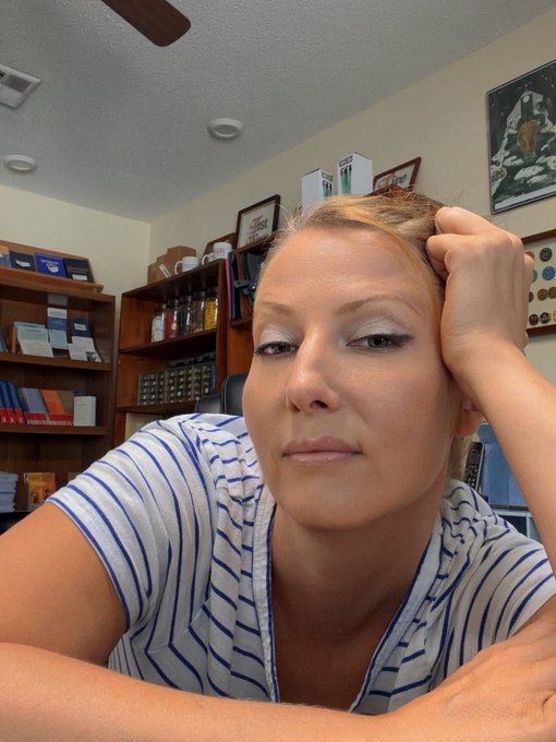 A woman with a contemplative expression rests her head on her hand in what appears to be an office or library setting, with bookshelves and framed pictures visible in the background, capturing a thoughtful moment likely during her volunteer work or registration for Artisan Cash.