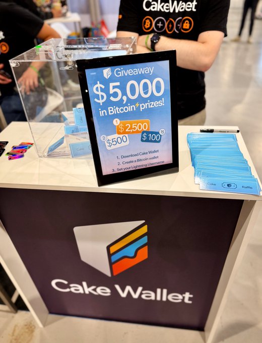 The image features a table at a convention or event displaying information about a bake sale, specifically offering $5,000 in Bitcoin prizes for anyone who participates. The cake wallet logo and an advertisement for the bake sale are prominently displayed on the table. A person is present in the scene but not interacting with the event itself. There are several other people visible at various locations within the image, contributing to a busy atmosphere surrounding the main table promoting the bake sale and Bitcoin prizes.