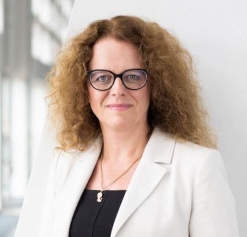 A professional headshot of a woman with curly blonde hair wearing glasses and a white blazer over a black top.