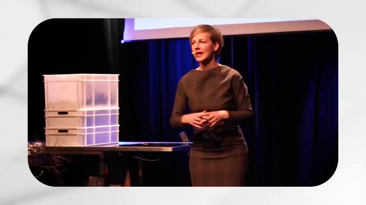 The image features a woman standing at a podium, giving a speech or presentation. She appears to be wearing a brown dress and has short hair. The background of the image shows a stage with a white curtain backdrop, suggesting that she might be speaking in front of an audience during a live event. There is also a small white box placed on the table near her, possibly containing props for her presentation or related to the content of her speech.