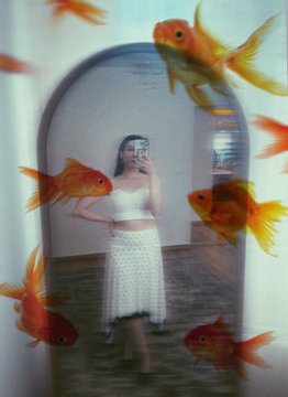 The image features a woman taking a selfie with her reflection visible through an archway or doorway, surrounded by goldfish swimming around her. The photo has been edited to create a surreal effect, making it appear as if she is standing inside the archway while the fish swim around her.