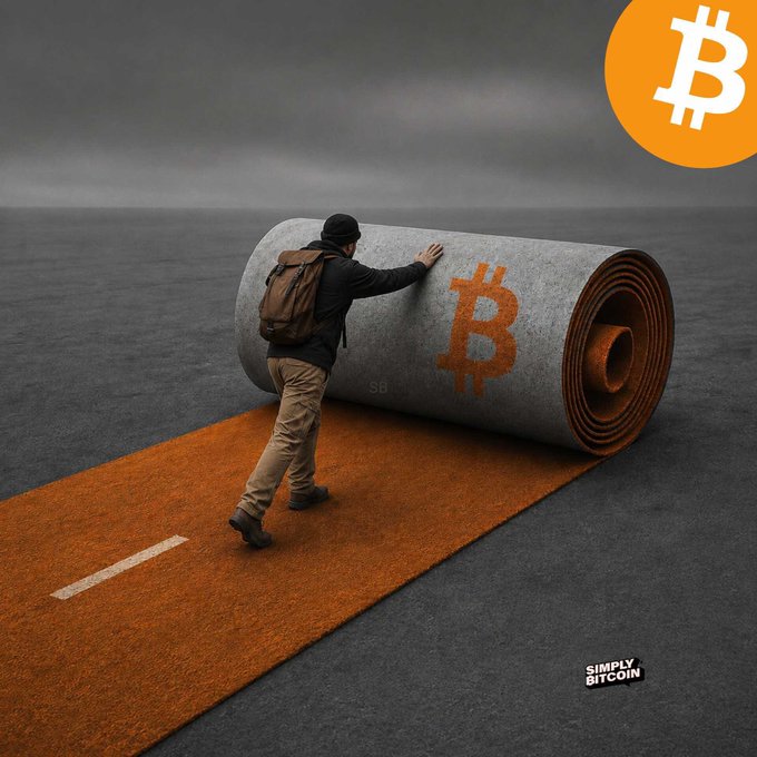 A man with a backpack pushes a large rolled-up carpet that unrolls to reveal an orange road featuring a Bitcoin symbol, with a Bitcoin logo in the corner and the watermark 'SIMPLY BITCOIN'.