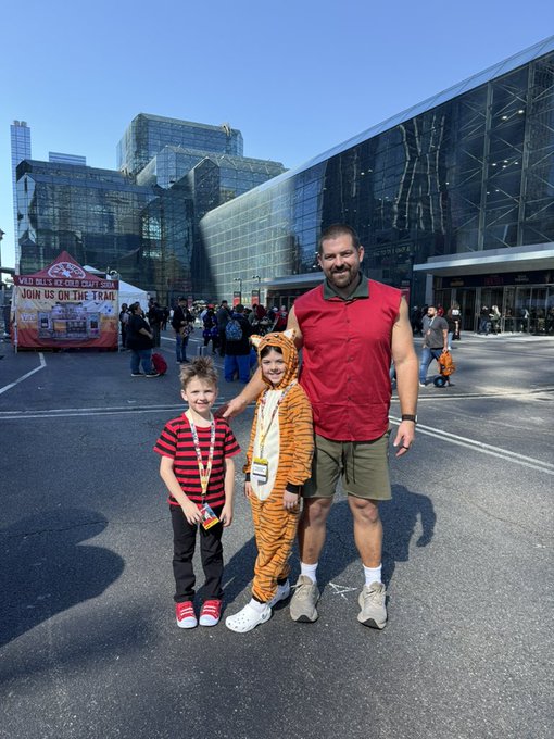 A man in a red sleeveless shirt and green shorts poses outside a convention center with two children, one of whom is dressed in a tiger costume and holding an event badge.