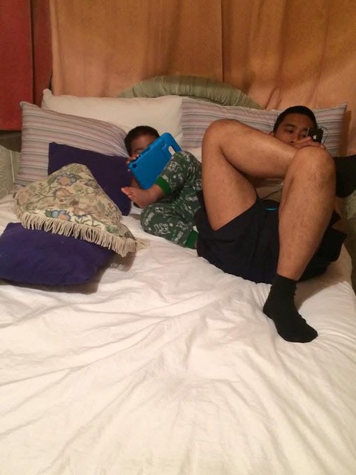 A person lies on a bed with their legs bent and crossed while a small child lies next to them playing with a blue tablet.