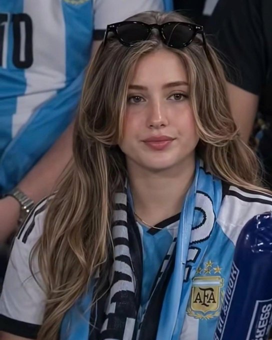 A young woman with long blonde hair and sunglasses perched on her head smiles softly while wearing an Argentina (AFA) soccer jersey, surrounded by fellow fans in a stadium setting. Her Argentine football attire mirrors the post's casual "bros" energy and weekend community vibe, adding a face to the laid-back, social-atmosphere message.
