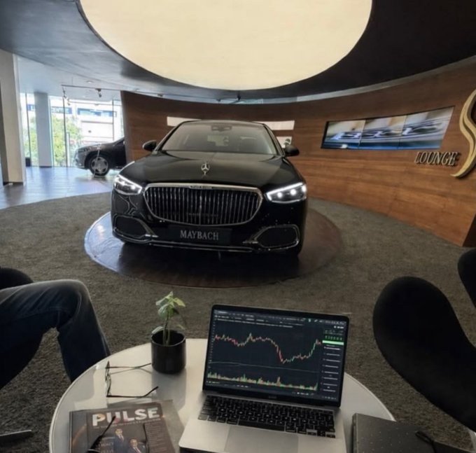 A Maybach luxury sedan is displayed in a sleek showroom "LOUNGE," while a laptop in the foreground shows a green upward-trending stock chart next to a magazine labeled "PULSE," visually reinforcing the post's "We're winning" sentiment through symbols of wealth and financial gains.