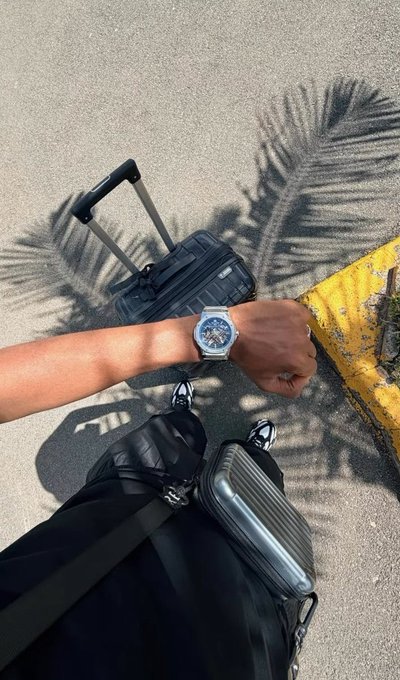 A traveler poses with their wristwatch and rolling suitcase on a sunny pavement, the shadow of a palm tree stretching across the scene—evoking a relaxed, intentional lifestyle. The image's travel and mindful-living aesthetic visually reinforces the post's message about blending mindfulness with crypto.