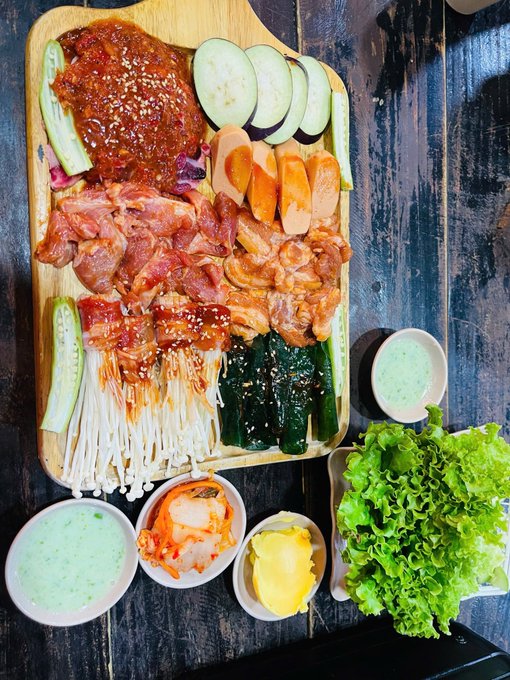 An overhead view of a wooden cutting board filled with raw meats including marinated beef and pork, enoki mushrooms, cucumbers, carrots, and lettuce, accompanied by dipping sauces.