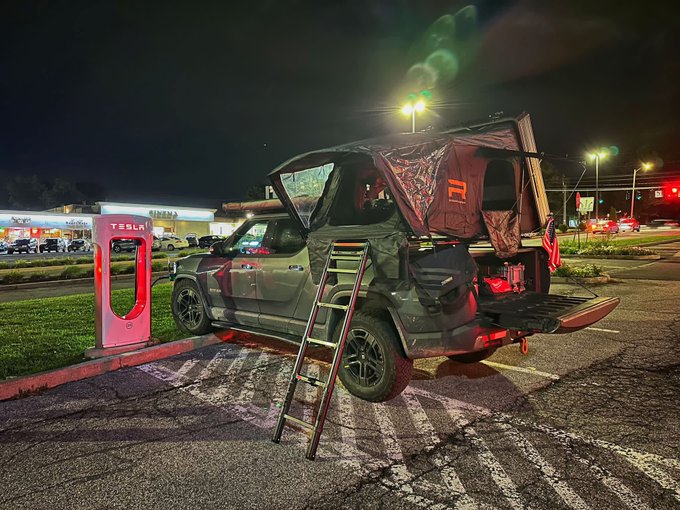 A Tesla Cybertruck outfitted with an overland camping setup including a rooftop tent and ladder, parked at a Tesla Supercharger station at night near a cinema.