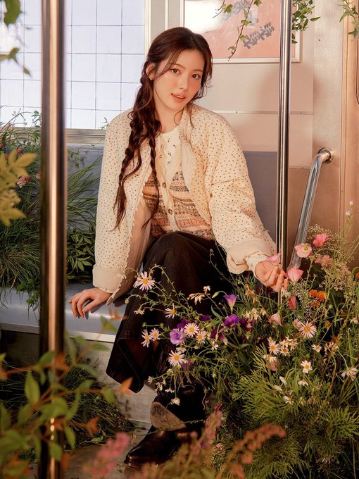 A young woman with long dark braided hair sitting on a subway or bus step, wearing a cream-colored jacket and patterned vest, surrounded by a lush arrangement of wildflowers.