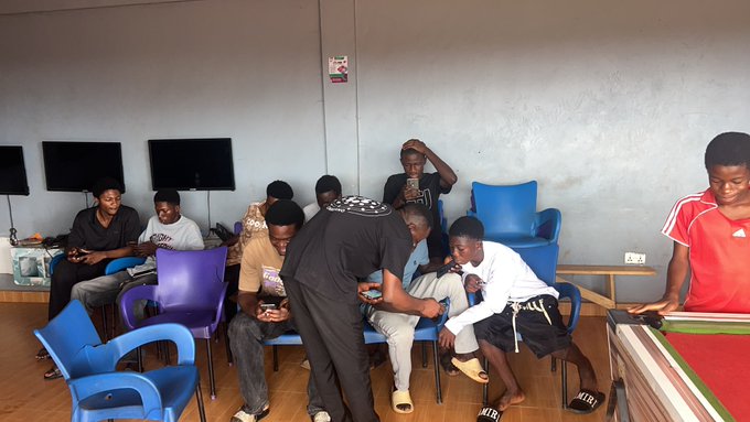 A group of young men gather in a room with blue chairs and computer monitors, where most are seated and looking at their phones while one stands and another plays pool on the right.