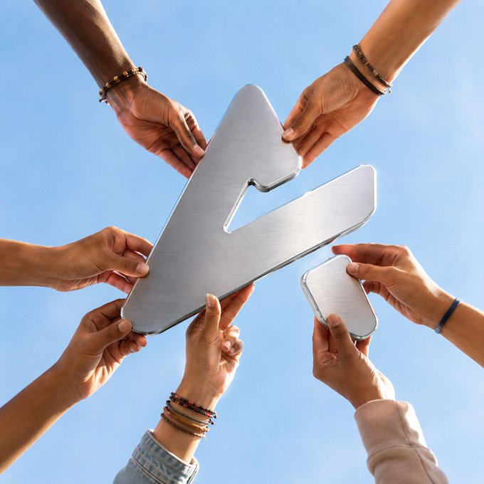Multiple hands of different skin tones and wearing various bracelets collectively hold up a large silver lightning bolt shape against a blue sky background.