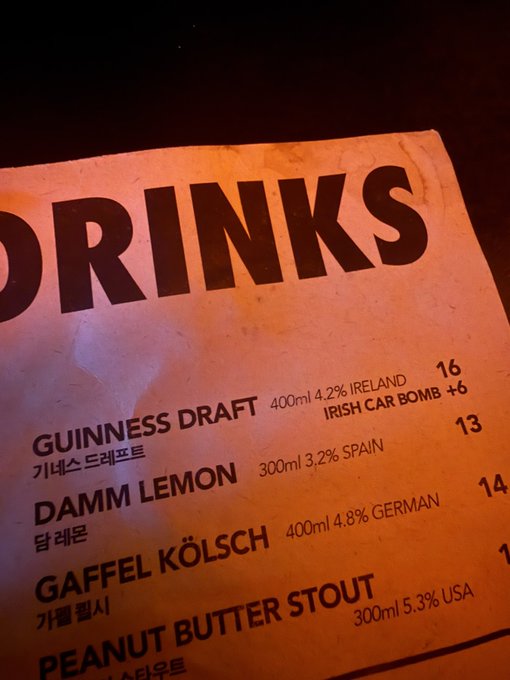 A drinks menu illuminated with warm light, listing options like Guinness Draft (400ml 4.2% Ireland, 16), Damm Lemon, Gaffel Kölsch, and Peanut Butter Stout with prices and Korean translations.