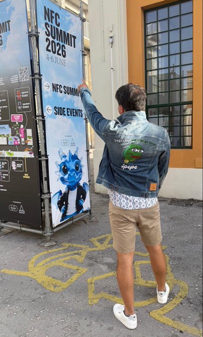 The image features a man wearing a denim jacket with a green frog design on the back pocket pointing towards an event poster for the NFCC Summit held at the NFCC Tower in 2016. The poster displays a blue cartoon frog, which appears to be the main attraction of the event. The man is standing outside and seems to be engaged in conversation or making plans related to the summit.