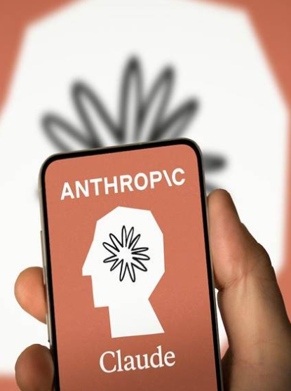 A hand holds a smartphone displaying the Anthropic logo featuring a head profile with a flower-like brain design and the text "ANTHROPIC" and "Claude" on a coral background, while a larger version of the same logo appears blurred behind it—visually branding the AI company associated with the tokenized ANTHROPICUSDT perpetual futures trading at valuations implying a $1.6T–$1.84T market cap.