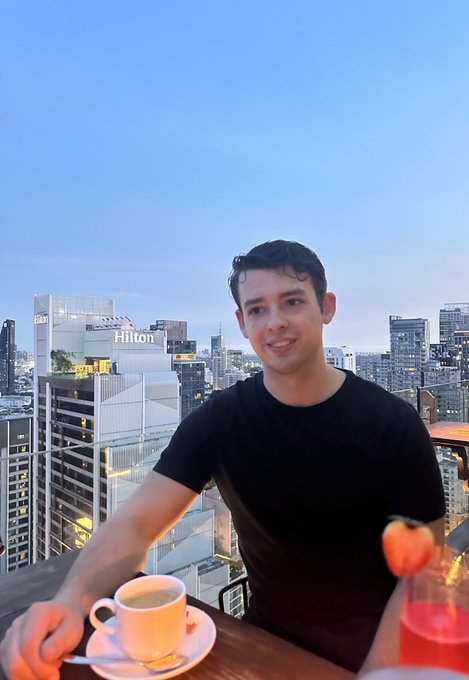 A young man in a black shirt smiles at the camera from a rooftop bar, with a coffee cup, a pink drink with a strawberry garnish, and a "Hilton" hotel building visible against the city skyline behind him—perfectly fitting the GM (good morning) vibe as he starts his day with a morning coffee and a friendly greeting.