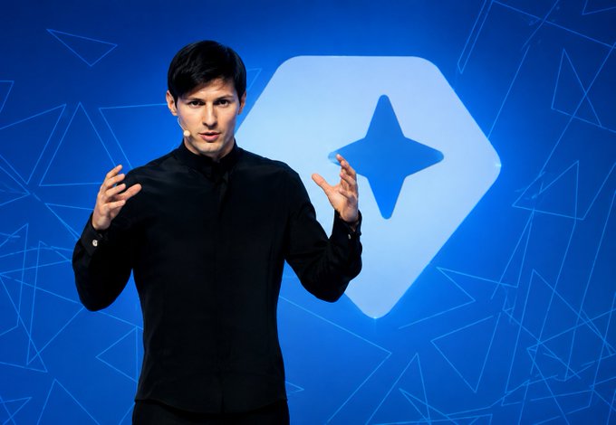 A man in a black shirt gestures expressively while speaking on stage, with a large white star/sparkle logo displayed on a blue geometric background behind him. The image likely depicts Pavel Durov, founder of Telegram and TON blockchain, in connection with the post's discussion comparing him to CZ (Binance) regarding ecosystem promotion.