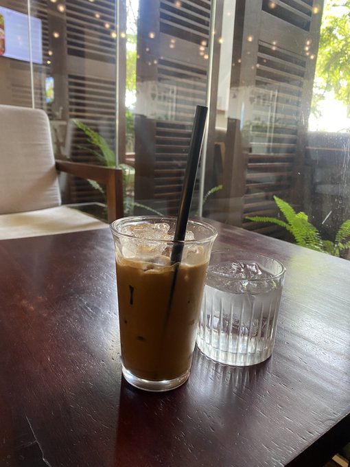 An iced coffee in a clear plastic cup with a black straw sits on a dark wooden table next to a glass of water, inside a cafe with large windows.