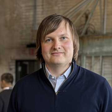 A man with light brown hair wearing a navy blue zip-up sweater over a light blue collared shirt stands smiling in what appears to be an industrial-style venue with exposed brick and metal trusses visible in the background. The post discusses Aave founder Stani Kulechov's criticism of proposed DeFi regulations from the ECB and EBA, suggesting the image likely depicts Kulechov himself voicing his concerns about the MiCA consultation responses.
