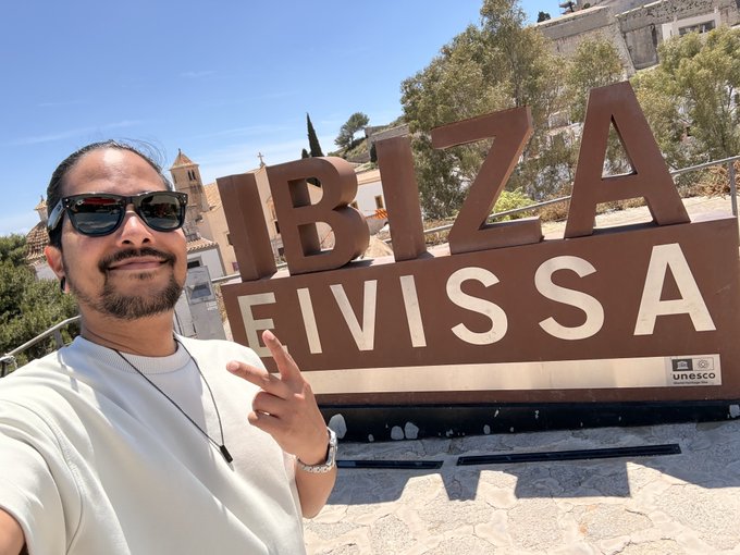 A man takes a selfie in front of the "IBIZA EIVISSA" sign with a UNESCO World Heritage logo visible, with stone architecture and trees in the background. The caption "Gm fam" suggests a casual morning greeting while sharing his travels at this iconic landmark.