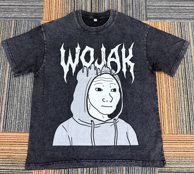 A black t-shirt featuring a graphic of a sad man wearing a hoodie and the text "Wojak" printed across the front. The t-shirt appears to be made of denim or rag material, with a corduroy texture. It is displayed on a patterned floor consisting of horizontal stripes in various colors including orange, yellow, and gray.