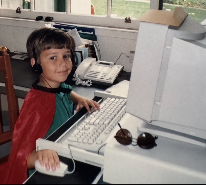 A vintage photograph of a young boy wearing headphones and a red cape, sitting at a desktop computer with a keyboard and mouse.