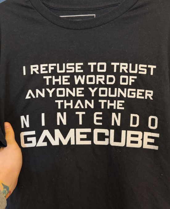 A black t-shirt held up displaying the text: 'I REFUSE TO TRUST THE WORD OF ANYONE YOUNGER THAN THE NINTENDO GAMECUBE'.